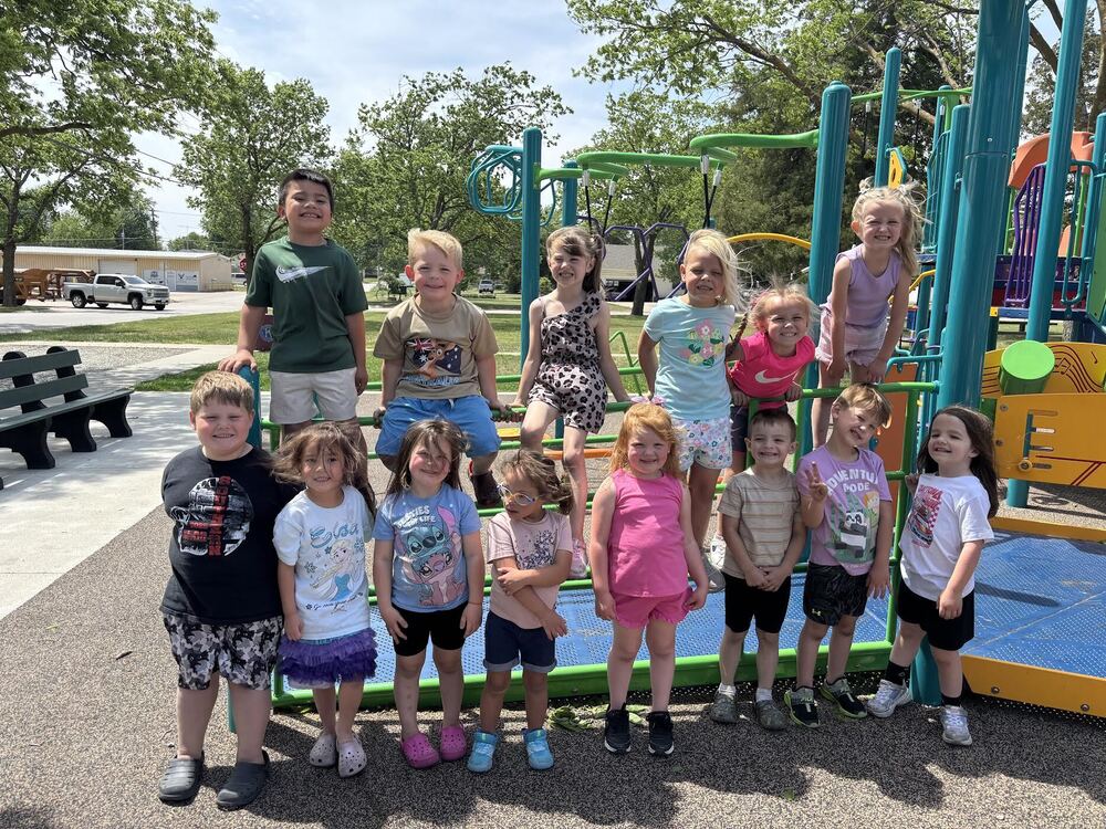 A group of fifteen young children pose together on a colorful playground structure at a park on a sunny day. Some children are standing on the ground in front, while others sit or stand on the climbing equipment behind them. Most are smiling and looking toward the camera. The playground features teal, green, yellow, and orange equipment surrounded by trees with green leaves. A bench, parked vehicles, and a small building are visible in the background. The sky is lightly clouded, and shadows on the ground suggest midday sunlight.