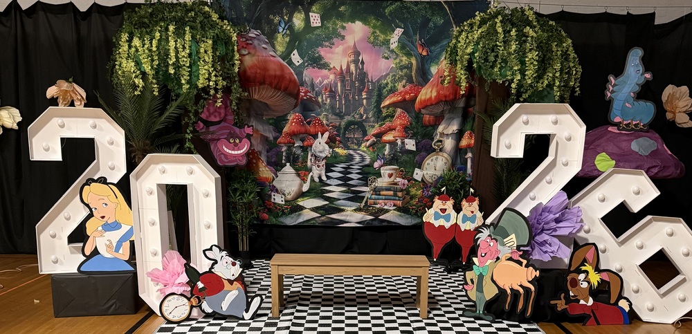 An Alice in Wonderland themed event display featuring large, illuminated marquee numbers for 2026. The setup includes a checkered floor and backdrop of a fantasy forest, surrounded by character cutouts like Alice, the White Rabbit, and the Mad Hatter.