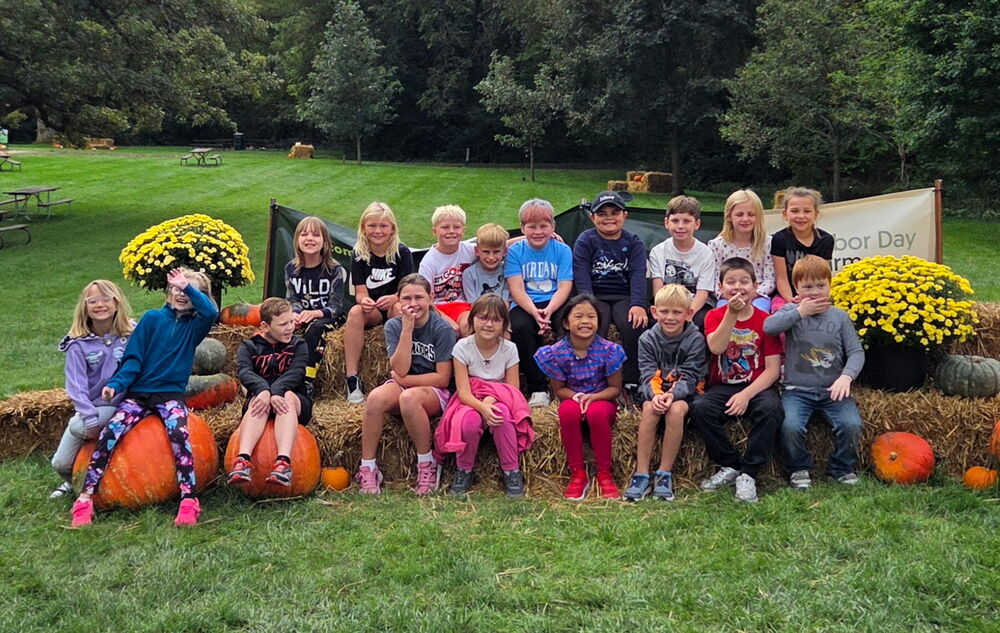 Students are sitting on hay bales with pumpkins and mums at their field trip to an apple orchard.