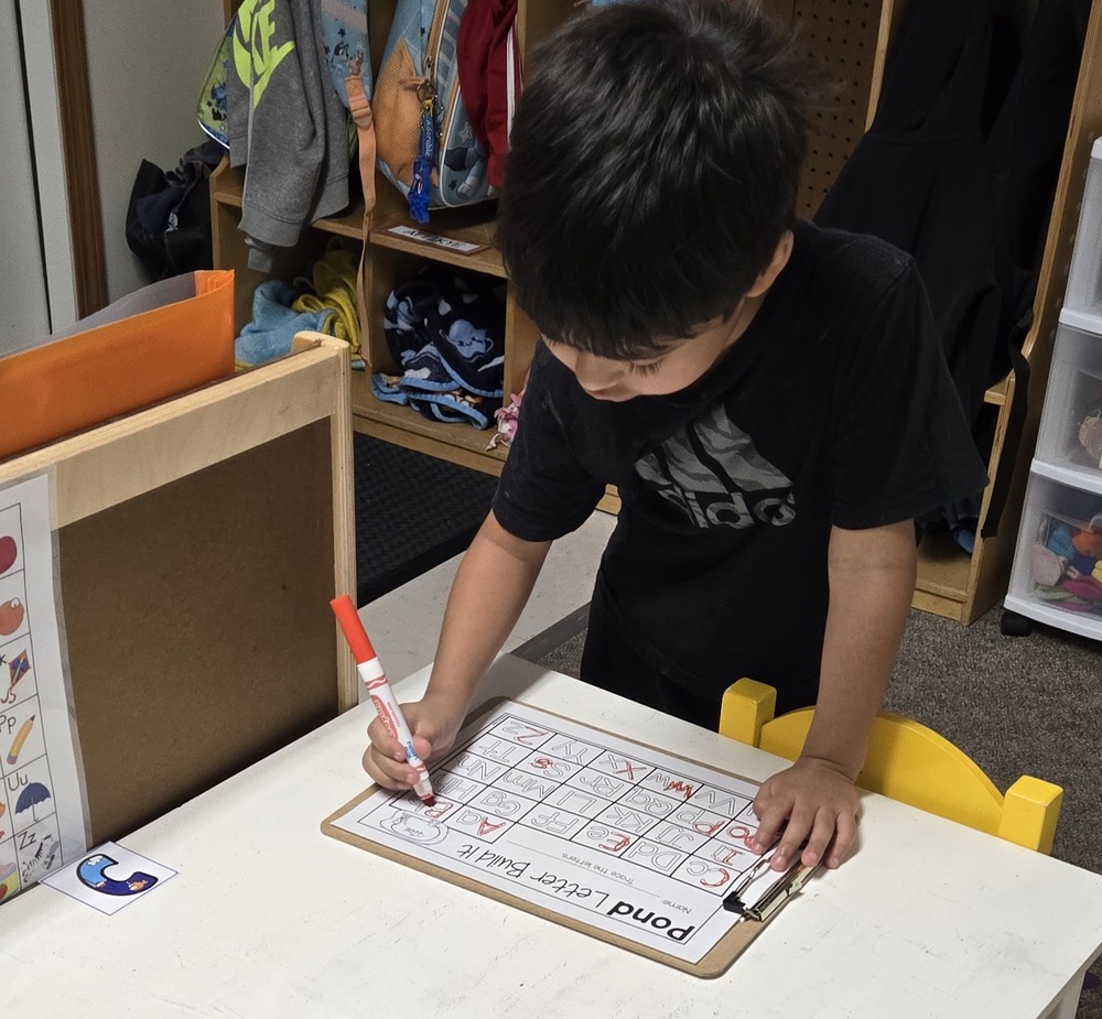 A young child stands at a small classroom table, using a red marker to complete a letter-building worksheet on a clipboard. The child is focused on tracing or circling letters, while cubbies with backpacks and clothing are visible in the background, creating a preschool or daycare classroom setting.