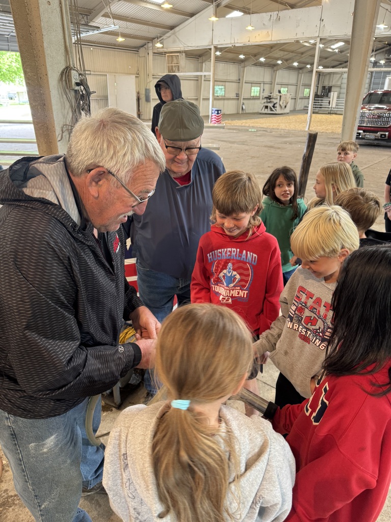 An older man demonstrates something with his hands to a small group of children gathered closely around him inside a barn. The children watch attentively. Another adult stands nearby observing.