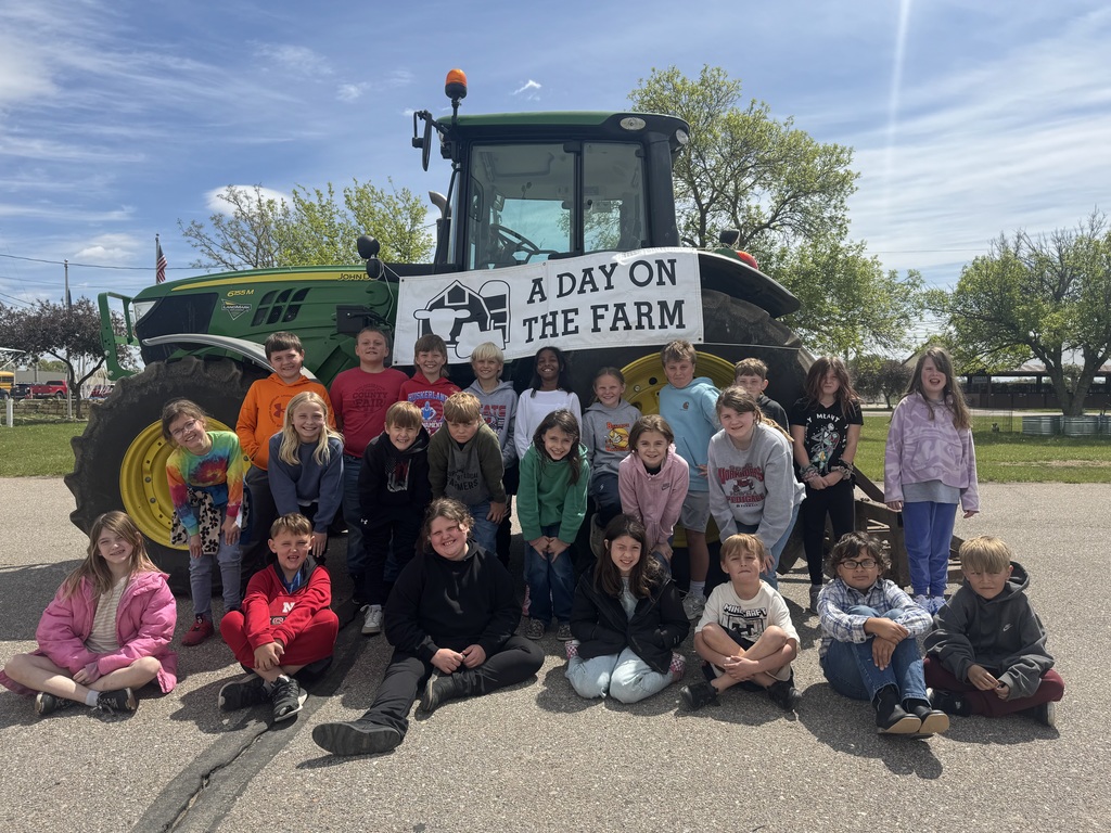 A group of children pose in front of a large green tractor outdoors. A sign on the tractor reads “A Day on the Farm.” The children smile and face the camera.