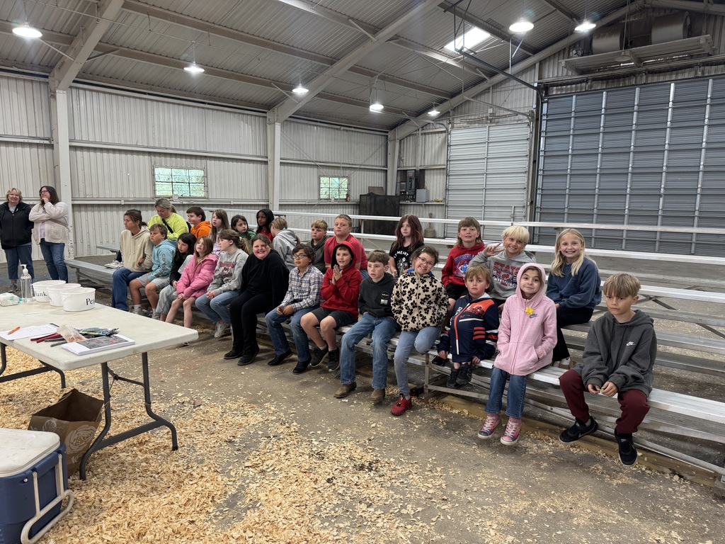 A large group of elementary-aged children sit on metal bleachers inside a barn-like building. A few adults stand to the left. The space has high ceilings, bright overhead lights, and wood shavings on the ground. A table with supplies is visible in the foreground.