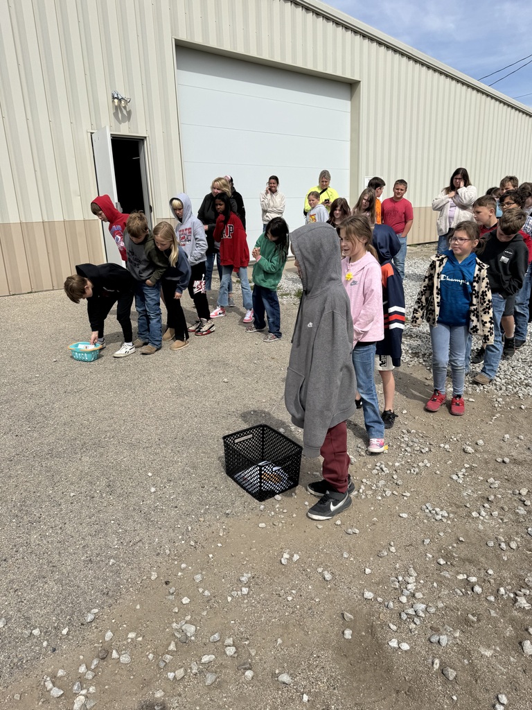 A group of children stand outside near a large building with a garage door. Some children are bending down to pick up items from the ground while others wait in line. Several adults stand in the background supervising.