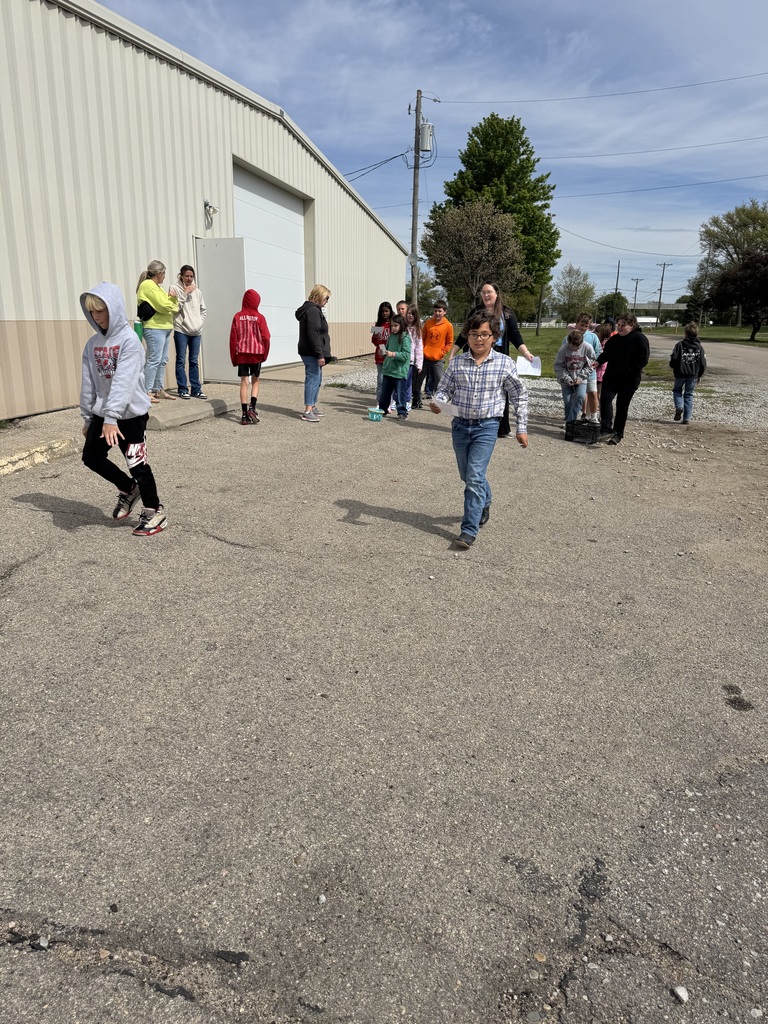 Children walk and gather outside near a large building on a sunny day. One child walks toward the camera while others move around in small groups. Adults stand nearby supervising the activity.