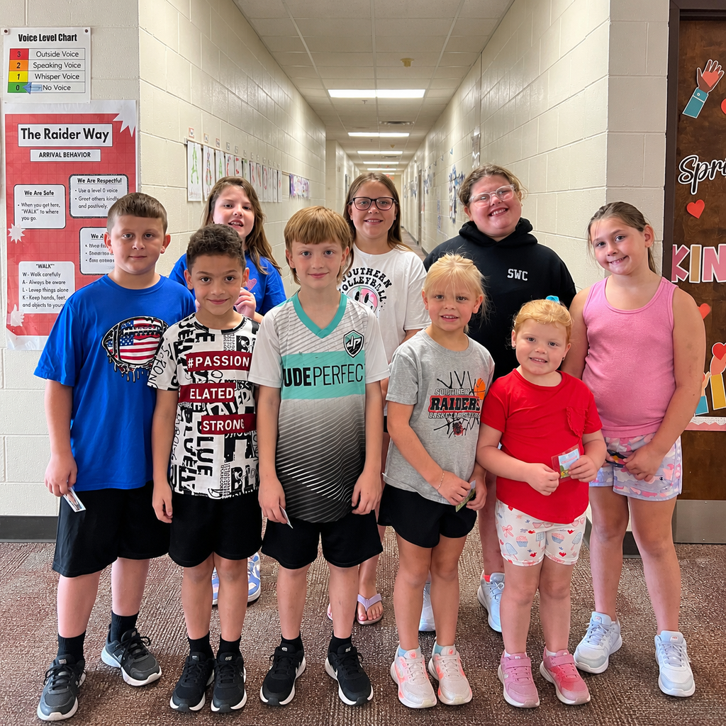 Eight Southern Elementary students pose together and smile for a photo in a school hallway. The students are arranged in two rows and are wearing colorful, casual school clothes. The hallway is decorated with student artwork, bulletin boards, and a red “The Raider Way” expectations poster.