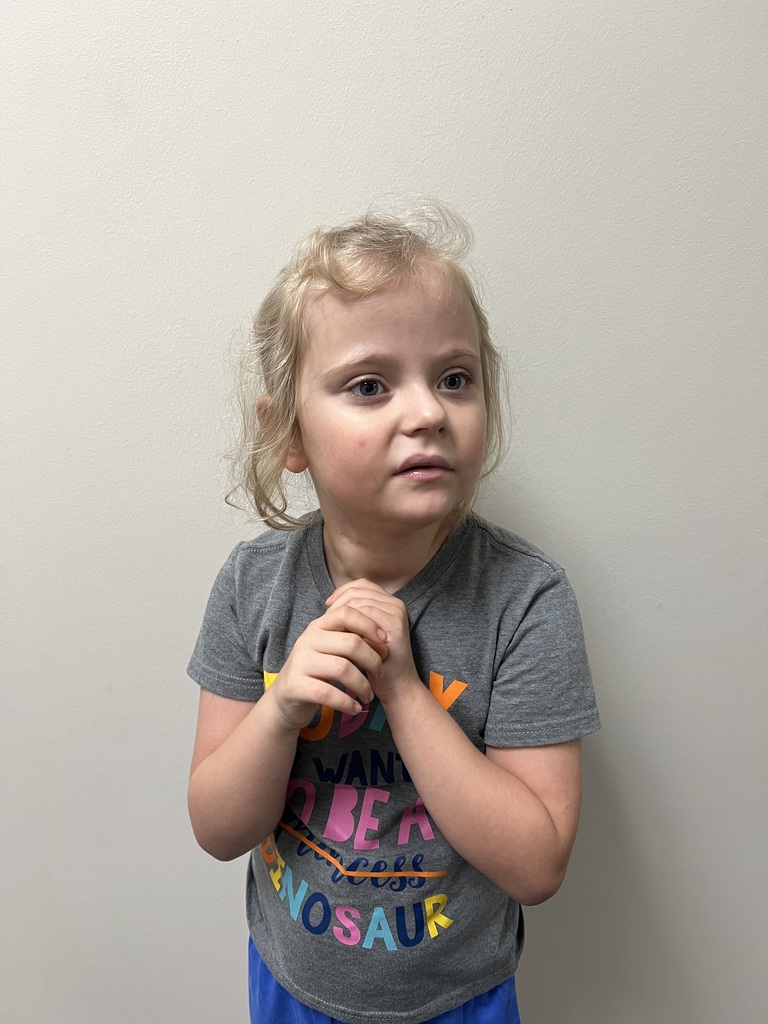 A young student with blonde hair stands against a light-colored wall with her hands clasped together in front of her. She is wearing a gray T-shirt with colorful lettering and blue bottoms and is looking slightly to the side of the camera.