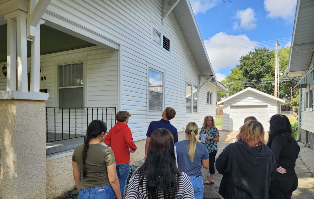 8 Students and Realtor looking at the outside of a vinyl sided 1920s style home to identify what to look for purchasing a home.