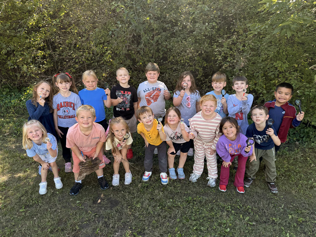 A group of kindergarten students smiling outdoors during a nature walk, holding magnifying glasses and other nature finds as they explore plants, trees, and their surroundings using their five senses.