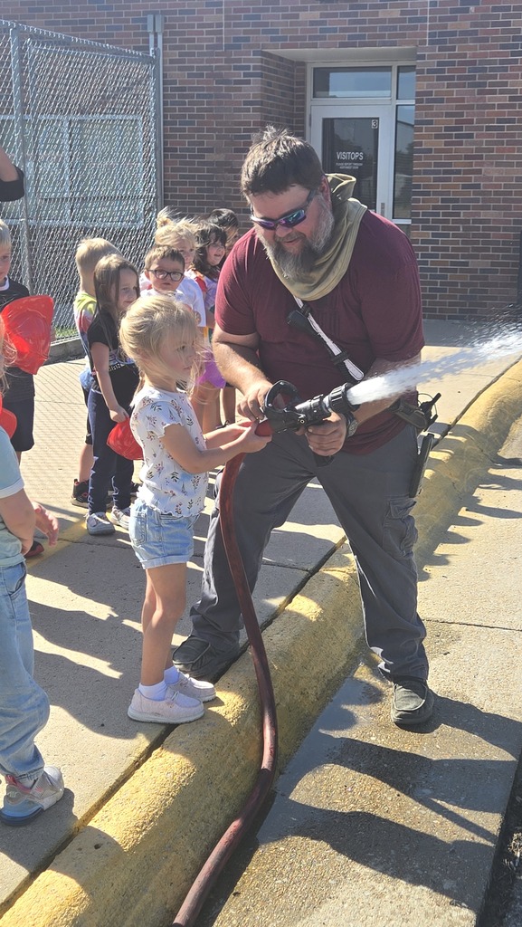 A young girl stands on a sunny sidewalk while a bearded man helps her hold a red fire hose that is spraying water. The girl, wearing a white patterned t-shirt and denim shorts, holds onto the hose near the nozzle. The man, dressed in a maroon t-shirt, sunglasses, and grey tactical pants, bends down to support the weight of the spraying hose. In the background, a line of other children stands along a brick school building with a window labeled "VISITORS," watching the activity.
