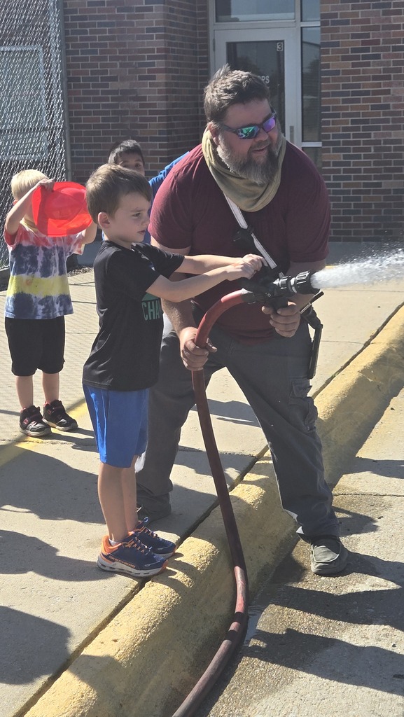 A young boy stands on a sunny sidewalk alongside a bearded man who is helping him hold and aim a spraying fire hose. The boy, wearing a black t-shirt and blue shorts, grips the nozzle with both hands while focusing intently on the stream of water shooting out. The man, wearing a maroon t-shirt, grey pants, and sunglasses, smiles warmly as he supports the heavy hose from behind. In the background, other children watch, including one wearing a tie-dye shirt holding a red plastic fire hat.