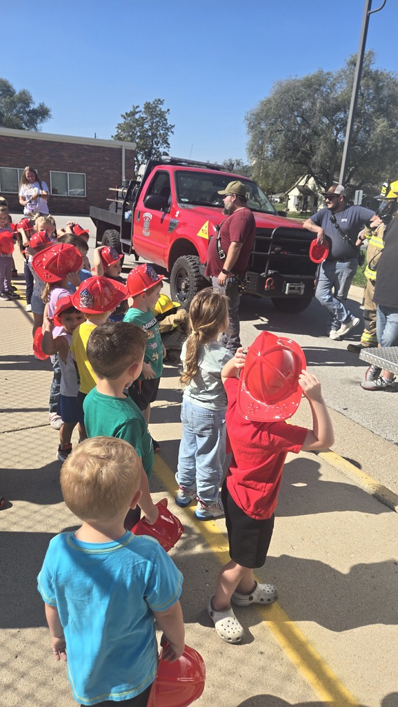 A group of young children wearing red plastic firefighter hats stand in a line on a sunny sidewalk, looking toward a red emergency truck and firefighters. The children, viewed mostly from behind and the side, are dressed in casual clothes like t-shirts, jeans, and shorts. In the background, a red utility truck is parked next to the curb, with two men in casual clothing and a firefighter in full turn-out gear interacting near the front of the vehicle. A brick building and green trees are visible under a clear blue sky.