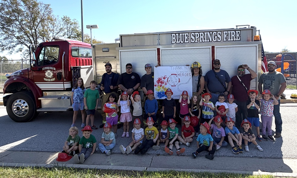A large group of children and firefighters pose for a photo in front of a red "Blue Springs Fire" engine on a sunny day. The children are arranged in two rows—some sitting on the curb in front and others standing behind them—with many wearing red plastic fire helmets. Several adults, including firefighters in uniform, stand behind the children. In the center, two individuals hold up a large, colorful hand-painted sign that reads, "Thank you to our heroes." An American flag is visible on the right side of the truck under a clear blue sky.