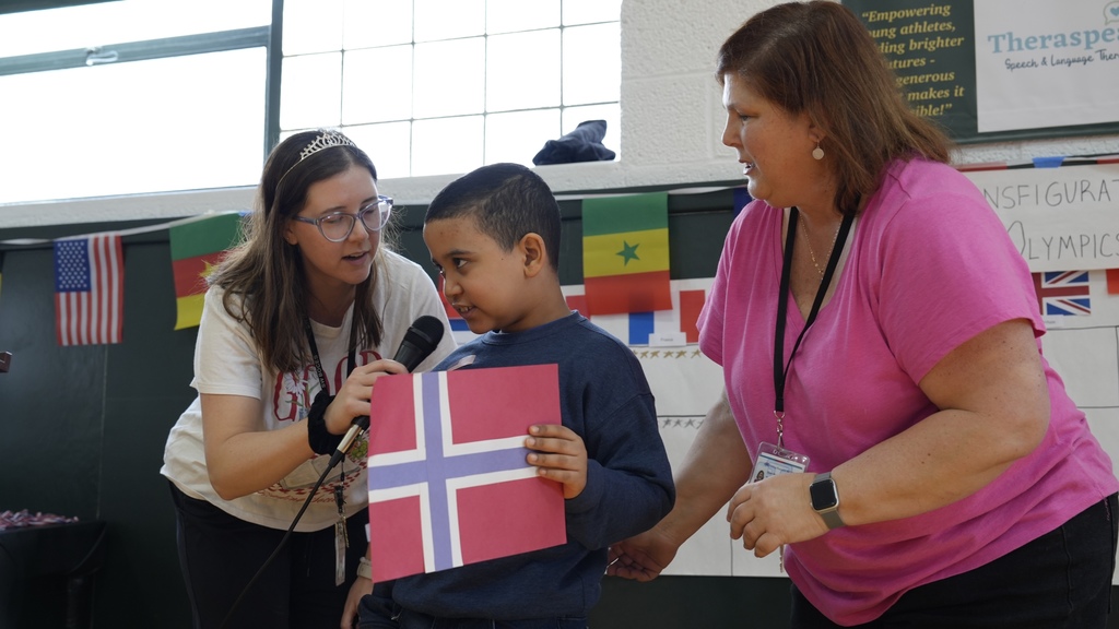 boy holds flag as teachers hold microphone to his face at event