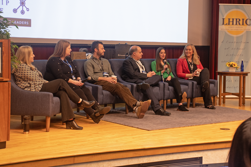 Six people sitting in blue chairs on a stage