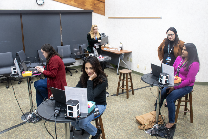 Four people sitting at tables with computers, one woman standing