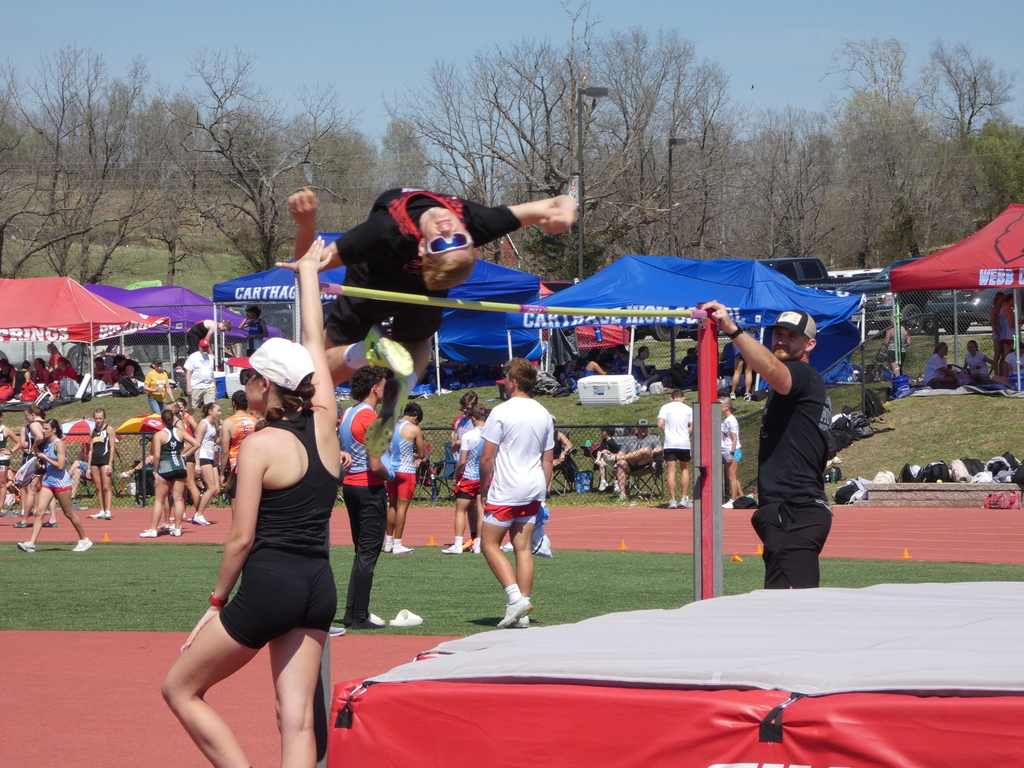An action-oriented photograph of a high jump competition at an outdoor track and field meet. The image captures a male athlete mid-air, successfully arching his back over a yellow crossbar. The Athlete: A young man wearing a black shirt with red accents, black shorts, and blue-tinted sunglasses is performing a "Fosbury Flop" high jump technique. His body is inverted and parallel to the ground as he clears the bar.  The Officials: Two individuals stand by the high jump standards:  On the left, a woman in a black tank top and white hat watches the jump with her hand raised.  On the right, a man in a black t-shirt and baseball cap stands near the upright post.  The Foreground: A large, thick red landing mat sits in the immediate foreground, ready for the athlete's landing.  The Background:  A red running track and green turf field are visible.  In the distance, several blue and red team tents are pitched on a grassy hill. One blue tent is labeled "Carthage High School" and a red tent is labeled "Webb City."  Numerous other student-athletes and spectators in various team uniforms are scattered across the field under a clear, bright sky.  Setting: The scene is a bright, sunny day at a well-attended high school track meet.