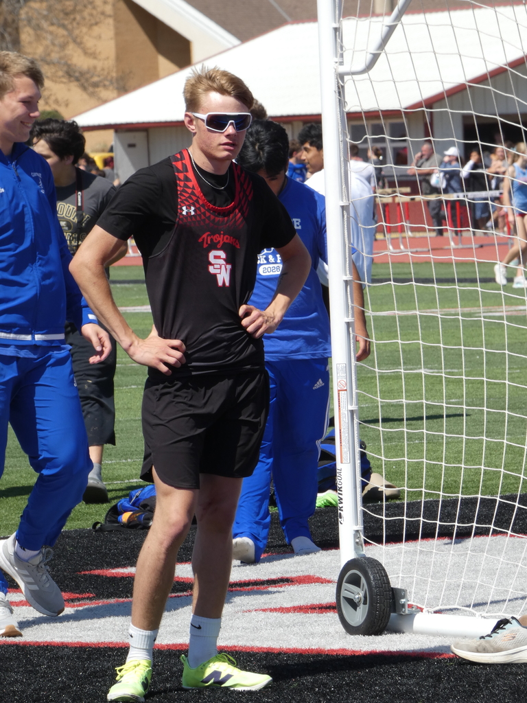 A full-length vertical photograph of a young male student-athlete standing on an outdoor sports field. He is dressed in track and field gear and stands with his hands on his hips, looking toward the left of the frame. The Subject: * The athlete has short blonde hair and is wearing white-framed, blue-tinted performance sunglasses.  He is wearing a black t-shirt underneath a black and red "Trojans" track singlet. The singlet features a red geometric pattern on the shoulders and the letters "SW" in red and white on the chest.  He is also wearing black athletic shorts, white crew socks, and bright neon yellow/green New Balance athletic shoes.  Other People: * To the left, another student in a blue track suit is partially visible.  Behind the main subject, other students in blue athletic wear are walking or standing on the field.  The Background:  The subject is standing on a black and white surfaced area of the field, near a white metal frame with a net (likely a soccer goal or similar sports equipment) on wheels.  In the far background, a red running track, white buildings with red trim, and hurdles are visible under bright, direct sunlight.