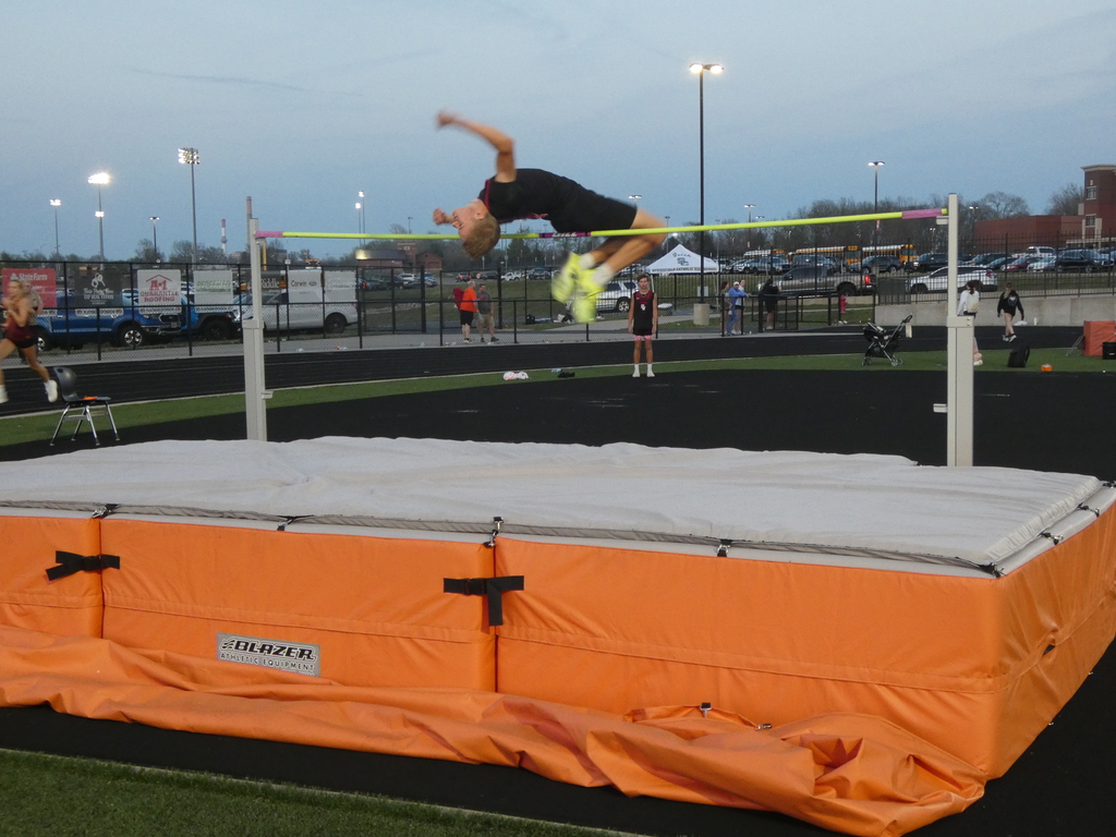 A wide-angle, landscape photograph of a high jump competition at dusk. The image captures a male student-athlete mid-flight, arching his body over a yellow crossbar during an evening track meet.