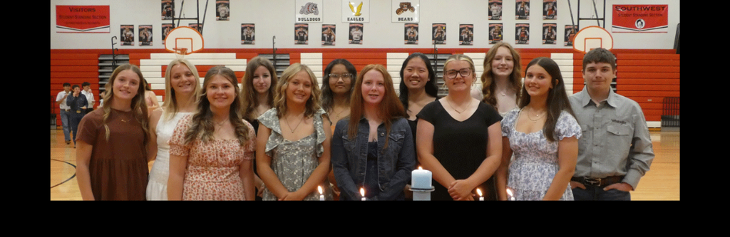 A horizontal group photograph featuring twelve students (eleven young women and one young man) posing for a formal portrait. The group is standing in a school gymnasium behind a ceremonial candle display, marking the 2026 National Honor Society (NHS) induction.