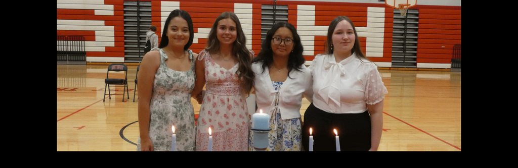 A horizontal group photograph of four young women standing side-by-side behind a candle-lighting display. They are posed in a school gymnasium for a National Honor Society (NHS) induction ceremony for 2026.