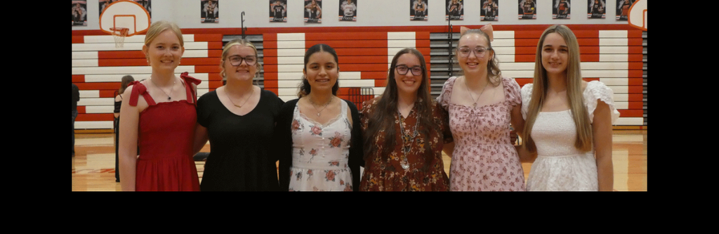 A horizontal group photograph of six young women standing in a line, posing for a formal portrait in a school gymnasium. This image is from a 2026 National Honor Society (NHS) induction ceremony