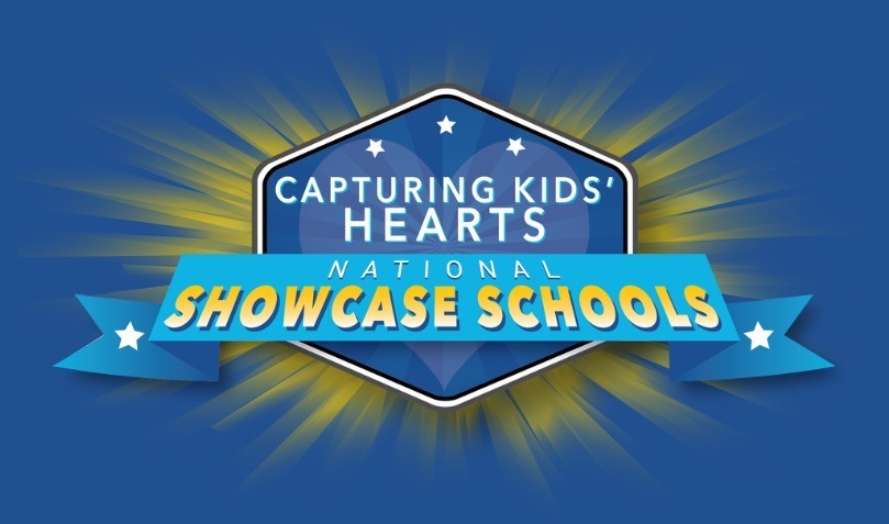 A graphic logo for Capturing Kids’ Hearts National Showcase Schools. It features a dark blue hexagonal shield with three white stars at the top and the text "CAPTURING KIDS' HEARTS" in white. A bright blue ribbon banner across the middle reads "NATIONAL SHOWCASE SCHOOLS" in bold yellow and white lettering. The emblem is set against a dark blue background with yellow light rays radiating from behind the shield.