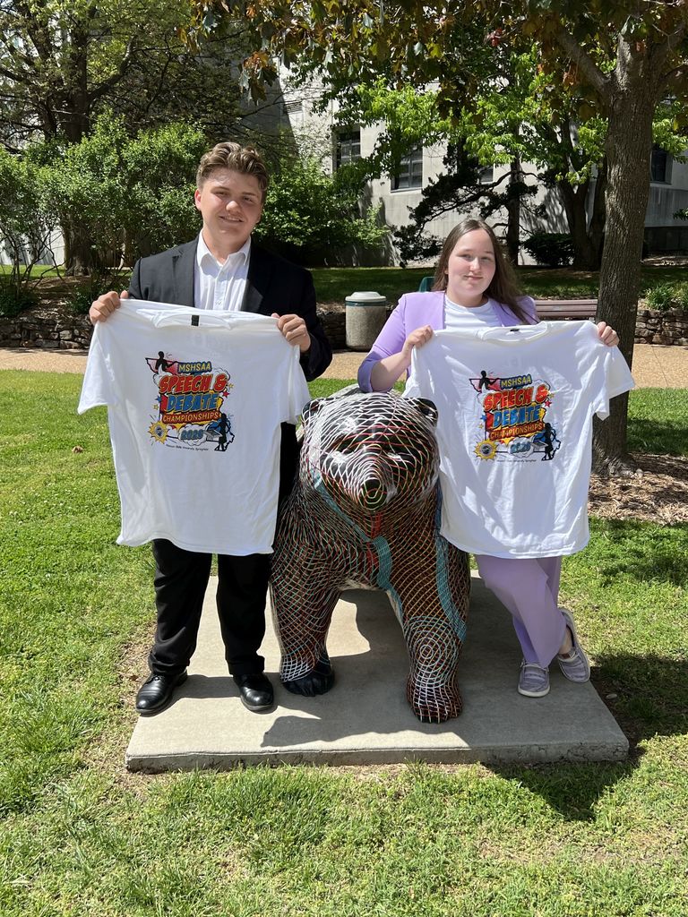 Here is the ADA-compliant alt text for the image:  **Alt Text:** A young man and a young woman stand outdoors on a sunny day, posing behind a decorative bear statue covered in a colorful, intricate wire-mesh pattern. The man, on the left, wears a black suit jacket and white shirt, while the woman, on the right, wears a lavender blazer and matching pants. They are both holding up white t-shirts featuring the "MSHSAA Speech & Debate Championships 2026" logo. The background shows a grassy area, trees, and a university building.