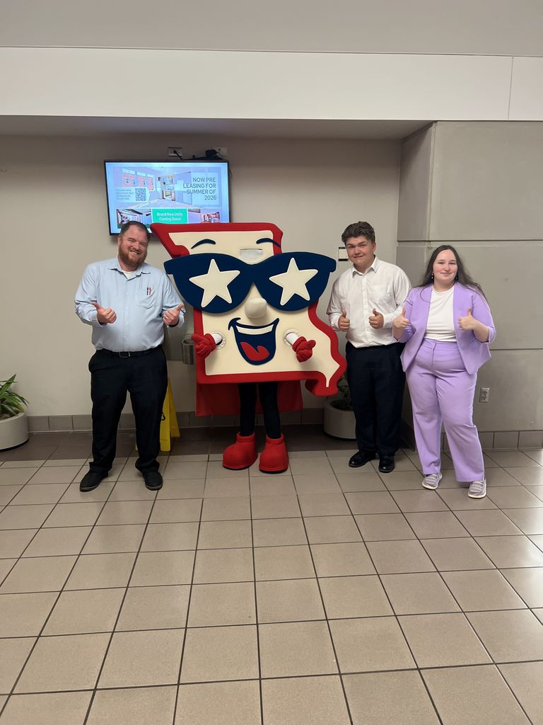 A photo of three people standing indoors next to a large mascot of the state of Missouri. To the left stands a man with a beard in a light blue button-down shirt and black slacks. To the right are a young man in a white shirt and black slacks, and a young woman in a lavender suit. All three individuals are smiling and giving a thumbs-up. The mascot in the center is shaped like Missouri, wearing large blue star-shaped sunglasses and a red cape.