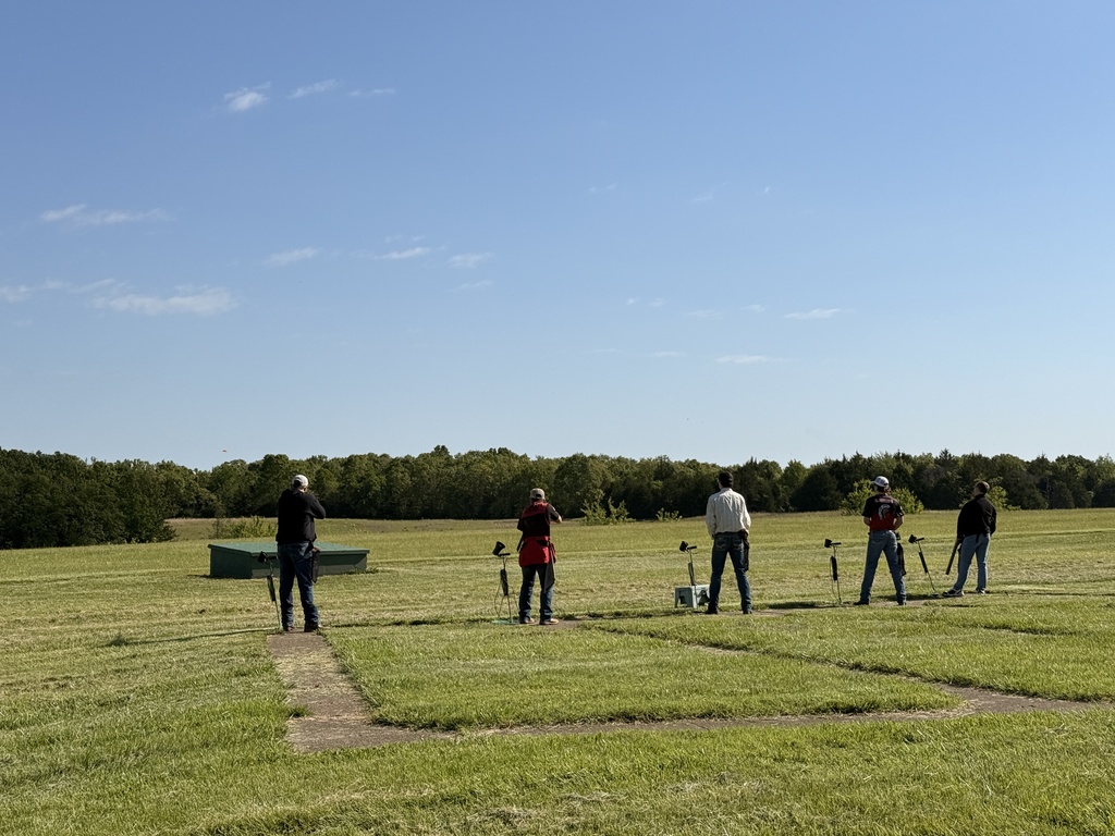 A wide outdoor shot of five individuals participating in trap shooting at a range. The shooters are spaced out along a concrete firing line on a large, green grassy field under a clear blue sky. Each person is seen from behind, facing toward a treeline in the distance. Small microphone stands and equipment cases are positioned near the shooters, and a low green bunker is visible in the field to the left.