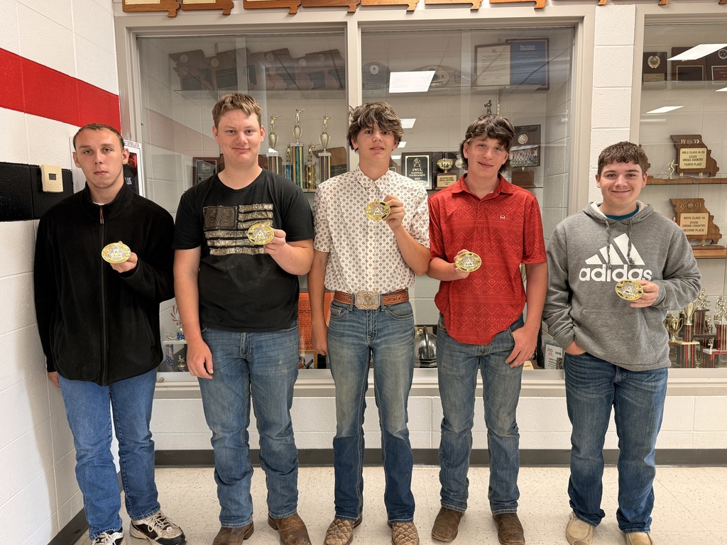 Five young men stand in a row inside a school hallway, posing in front of a glass trophy case filled with awards. Each individual is holding up a circular gold award buckle toward the camera. They are dressed casually in t-shirts, polos, or hoodies with jeans. The school wall to the left features a bold red and black stripe design.