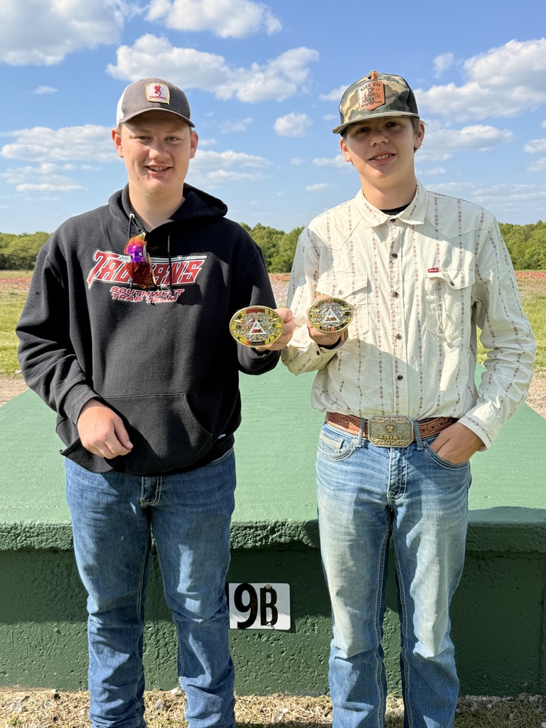 Two young men stand side-by-side outdoors on a sunny day, holding up award belt buckles. The man on the left wears a grey baseball cap, sunglasses tucked into a black "Southwest Trap Team Trojans" hoodie, and blue jeans. The man on the right wears a camouflage baseball cap, a light-colored patterned button-down shirt, and blue jeans with a large "Ariat" belt buckle. They are standing in front of a green concrete structure labeled "9B," with a grassy field and a line of trees in the background under a blue sky with light clouds.