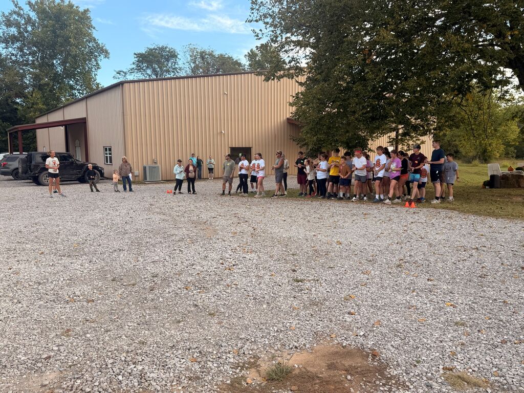 Here is the ADA-compliant alt text for the file "IMG_7396.jpg": **Alt Text:** A wide outdoor photo showing a group of students and adults gathered on a large gravel lot before a race or outdoor event. A large, beige metal building stands in the background alongside mature green trees under a partly cloudy sky. To the left, an event leader stands apart addressing the crowd near parked vehicles, while two small orange cones mark a starting area on the gravel near the crowd on the right.