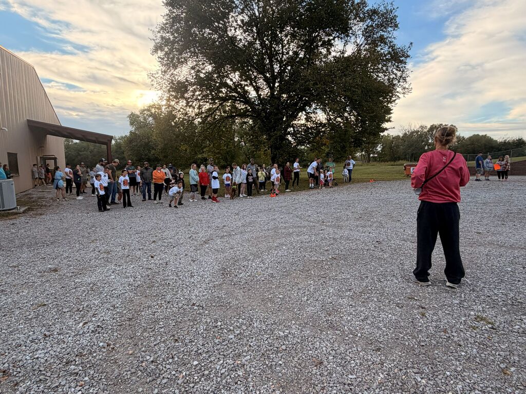 Here is the ADA-compliant alt text for the file "IMG_7392.jpg": **Alt Text:** An outdoor wide-angle shot from behind a speaker wearing a long-sleeved pink shirt and black pants, who is addressing a large crowd of adults and children gathered across a gravel lot. In the background, participants stand lined up near the side of a beige building under a large shade tree, with the sun low in a cloudy sky.