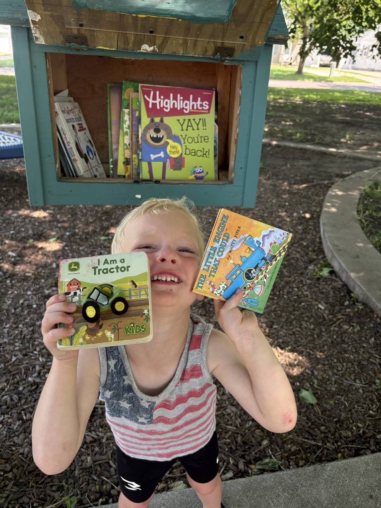 Young child smiling with books in front of a little library.