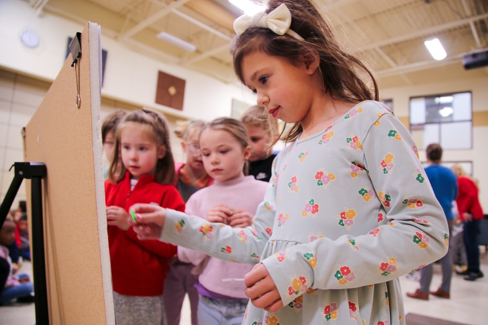 Young girl places a green sticker on a posterboard as classmates line up beside her.