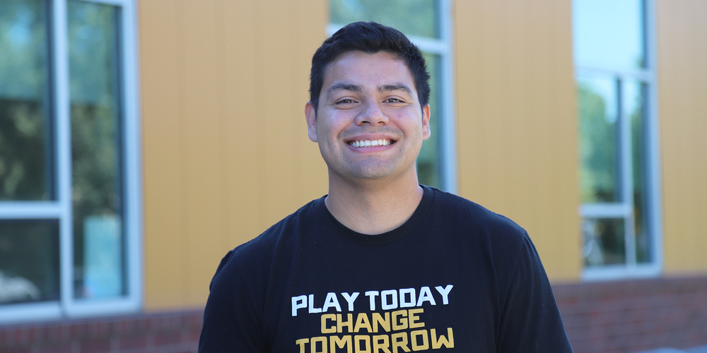 The image shows Austin Johnson smiling and standing in front of a building with large windows. He is wearing a black T-shirt that reads "Play Today Change Tomorrow" in bold white and yellow letters. The background is slightly blurred, showing a modern structure with mustard yellow walls and reflective glass panels.