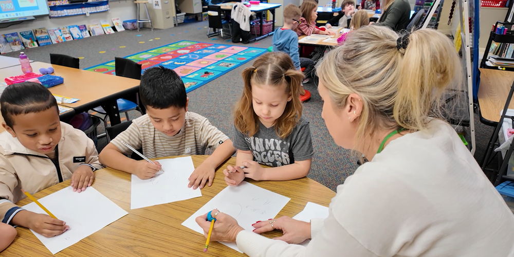 A teacher sits with kindergarten student at a table, holding pencils and practices handwriting.