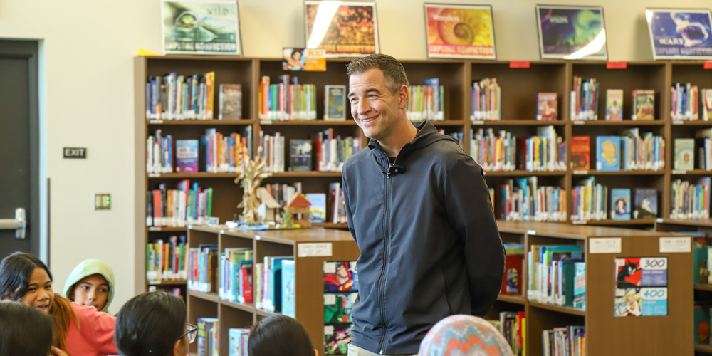 Superintendent Dr. Adam Swinyard smiles while standing in front of a group of students in a school library.
