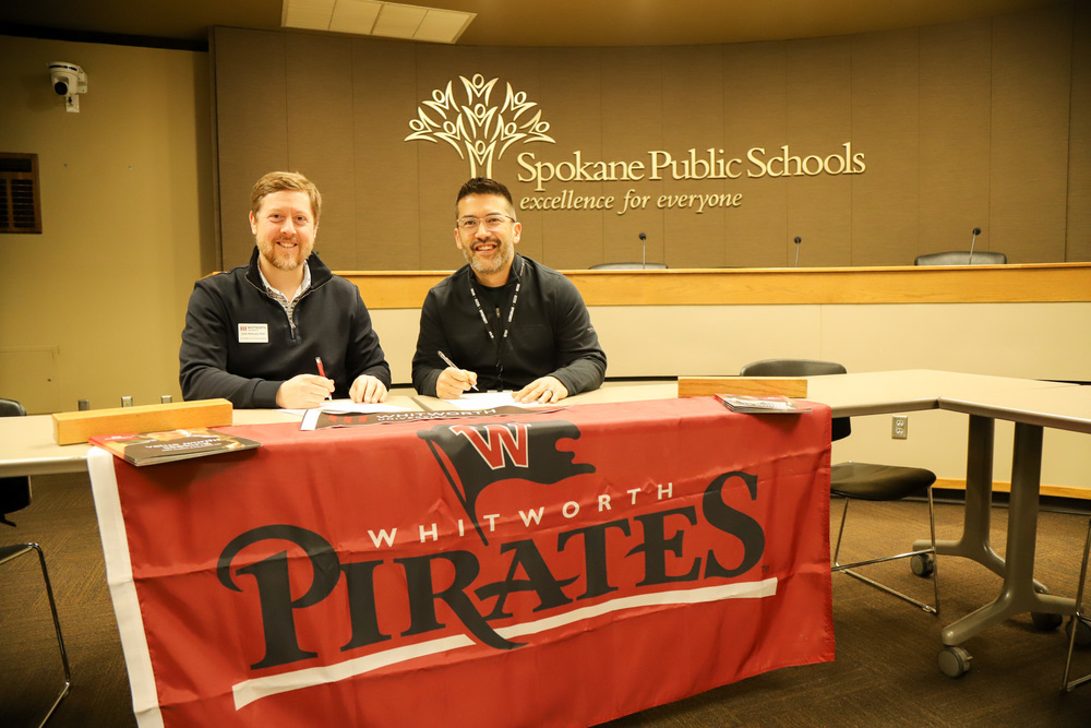 Josh Hibbard and Scott Kerwien smile at a desk with a flag for Whitworth Pirates; the Spokane Public Schools logo is displayed on the wall behind them.