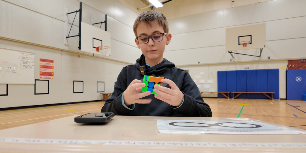 A student solves a Rubik's Cube while seated at a table in an elementary school gym.