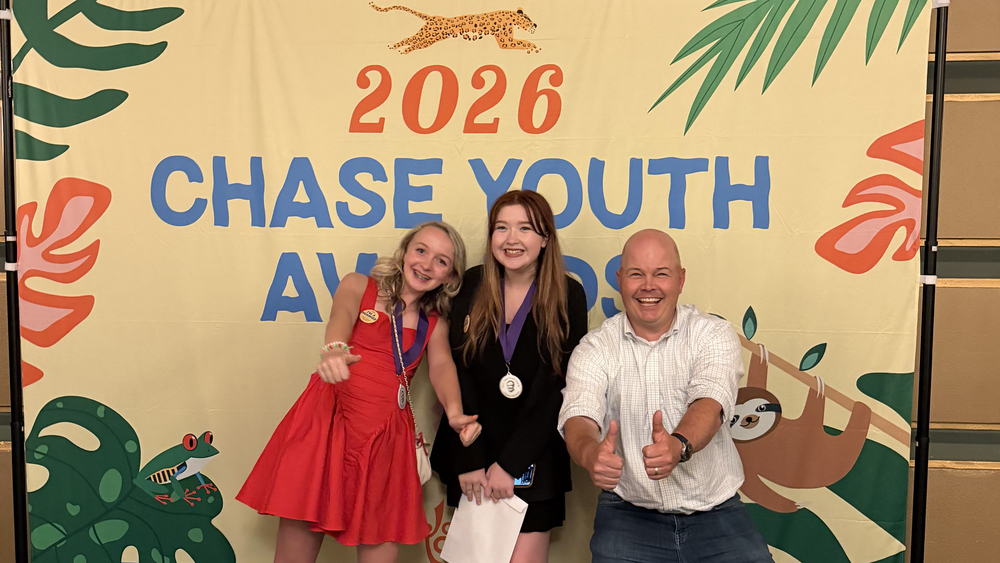 Two students wearing medals and an adult smile while giving thumbs up next to a banner for 2026 Chase Youth Awards.