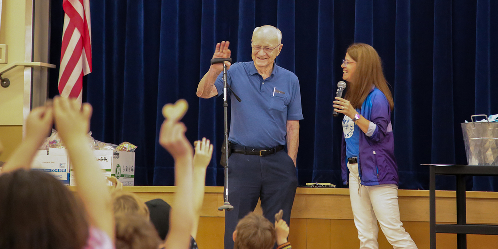 Lanny Burrill smiles and waves with Anne Walter as elementary students hold up wooden hearts during an assembly.