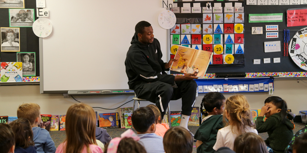 A college basketball player reads a book to elementary students in a classroom.