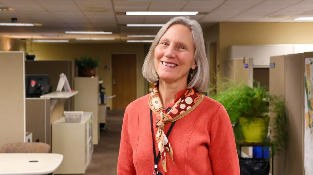Carol Pederson stands in an office, smiling, wearing orange sweater.