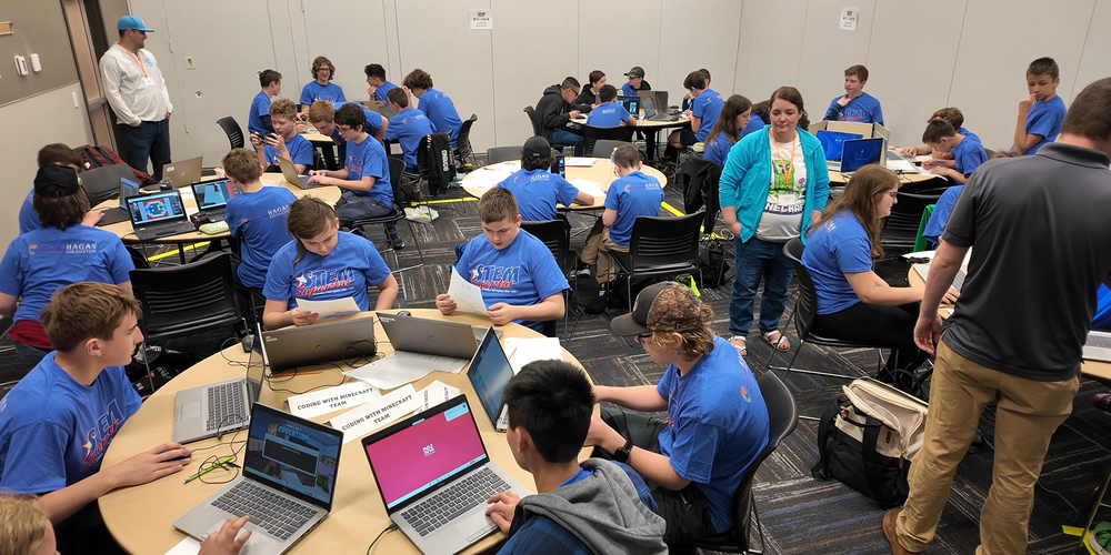 Middle school students seated at tables with laptops for a coding competition.