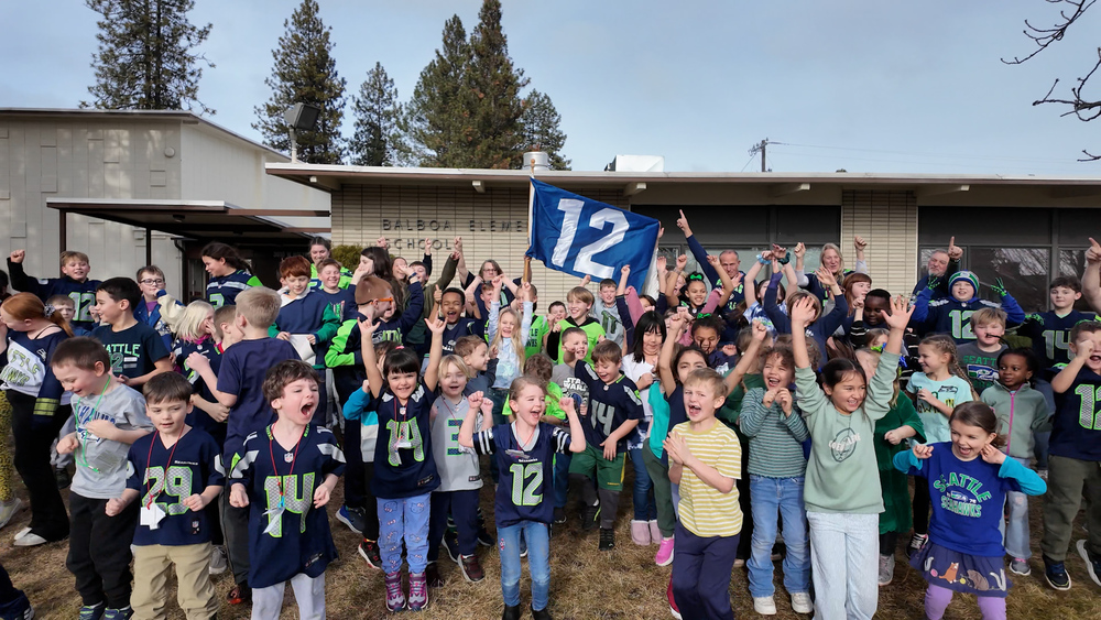 A large group of elementary students and staff wearing blue and green Seattle Seahawks cheer in front of a school.