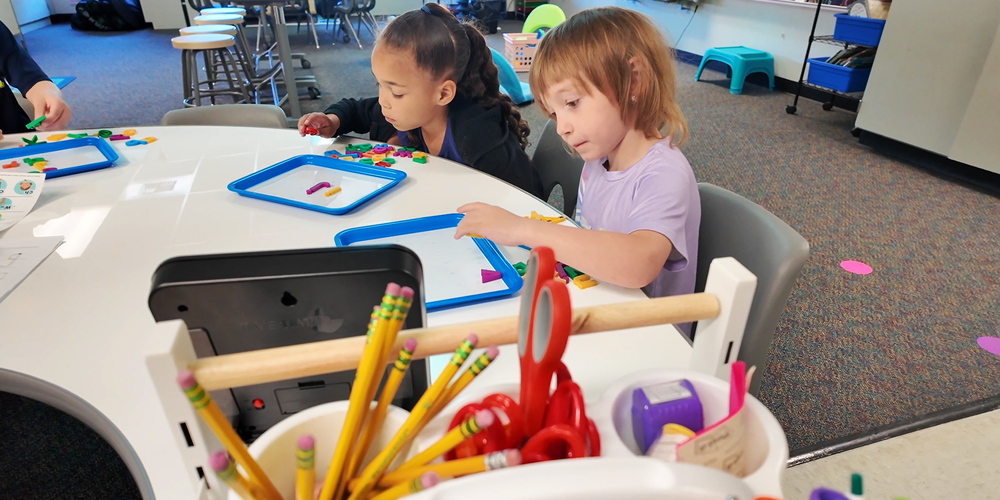 Two kindergarten students work with magnetic letters at a table.