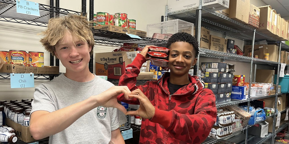 Two Salk Middle School students make a heart with their hands while standing in a food pantry.