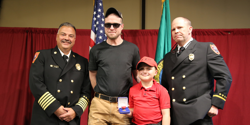 An elementary student smiles while holding a medal with his dad and two firefighters.